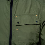 Close-up of an olive green jacket featuring a black zipper, a chest pocket with black snap buttons, and copper-colored rivets. MA.Strum Jacket available at Sakks Menswear in Southampton.
