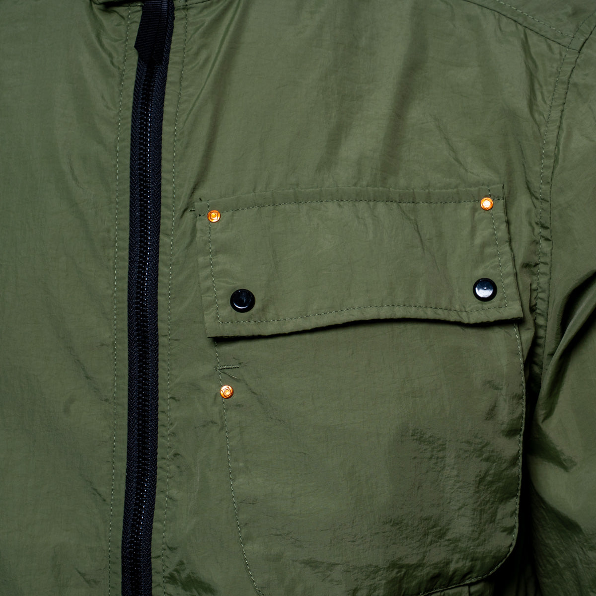 Close-up of an olive green jacket featuring a black zipper, a chest pocket with black snap buttons, and copper-colored rivets. MA.Strum Jacket available at Sakks Menswear in Southampton.