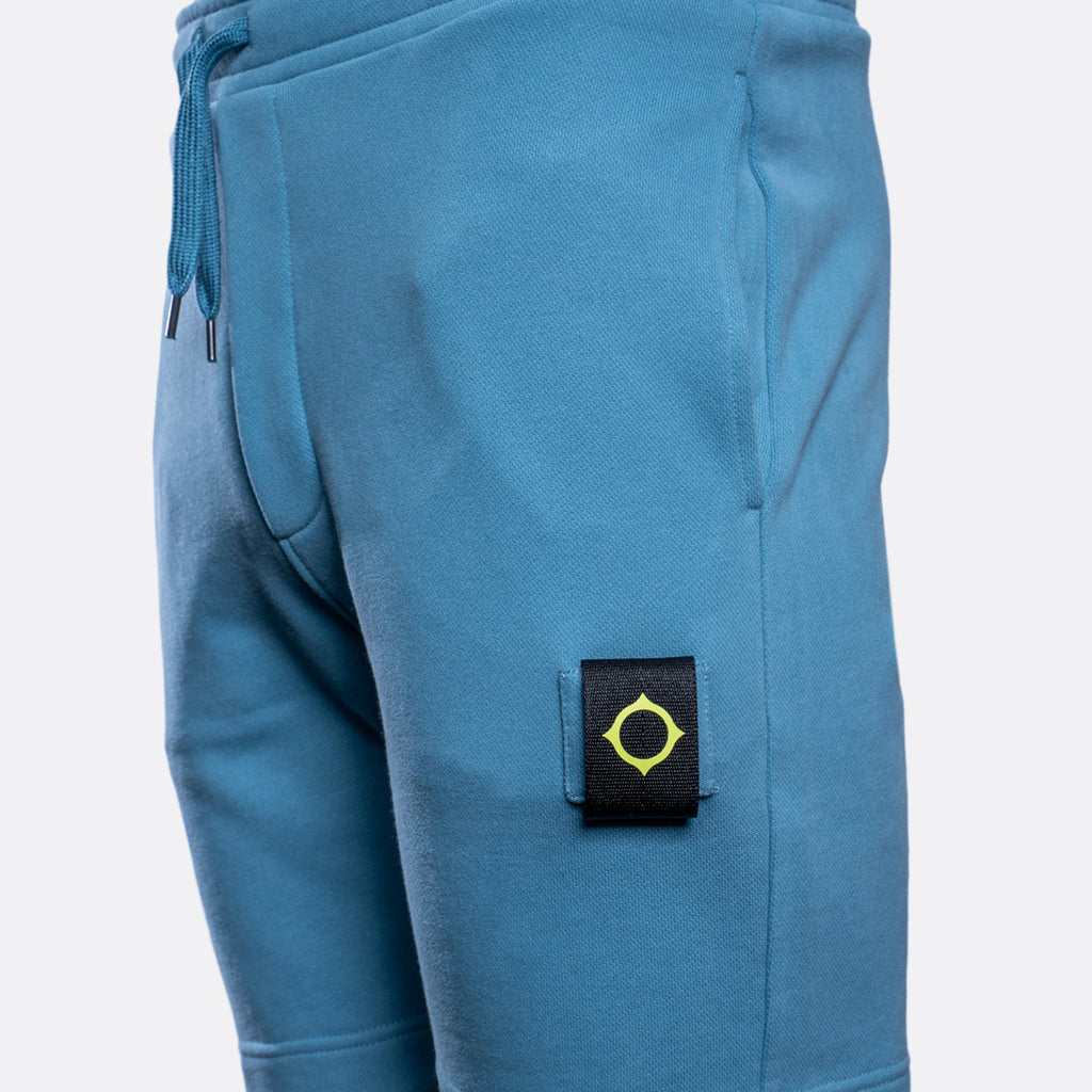 Close-up of a pair of blue sweatpants showing the side pocket with a black brand patch featuring a yellow compass logo, on a light gray background. MA.Strum Sweat Shorts available at Sakks Menswear in Southampton.