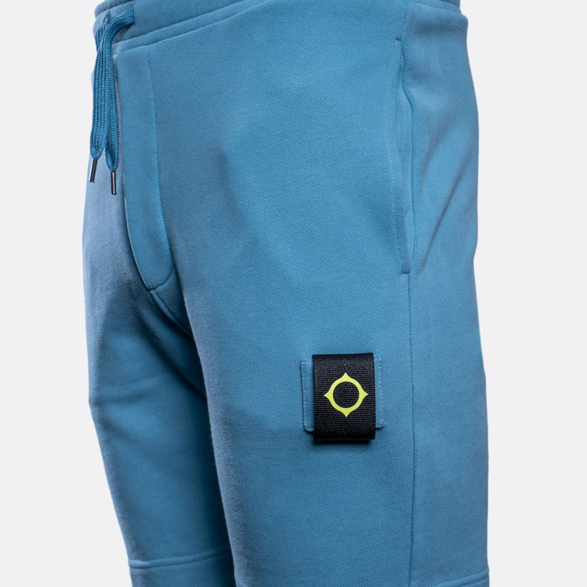 Close-up of a pair of blue sweatpants showing the side pocket with a black brand patch featuring a yellow compass logo, on a light gray background. MA.Strum Sweat Shorts available at Sakks Menswear in Southampton.