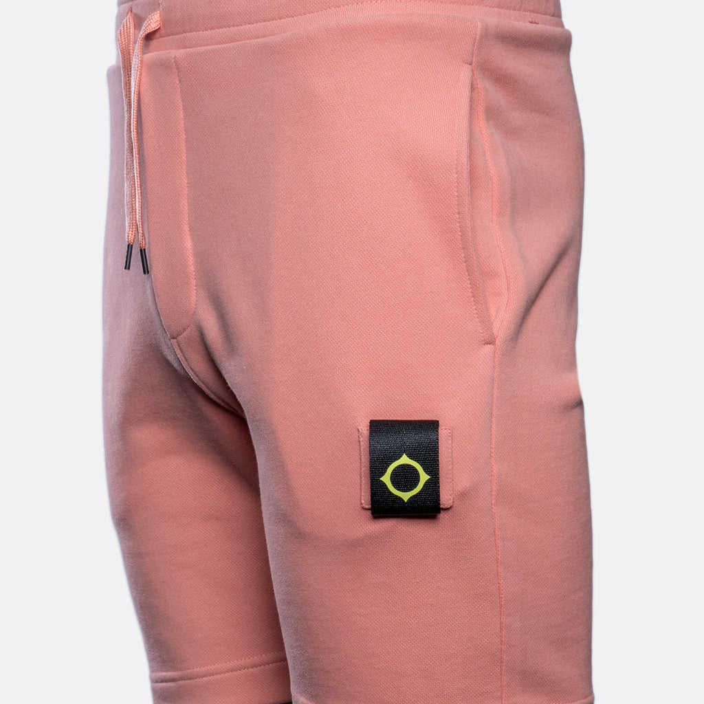 Close-up of a pair of dusty pink sweatpants featuring a black brand patch with a yellow-green compass logo on the upper thigh. MA.Strum Sweat Shorts available at Sakks Menswear in Southampton.