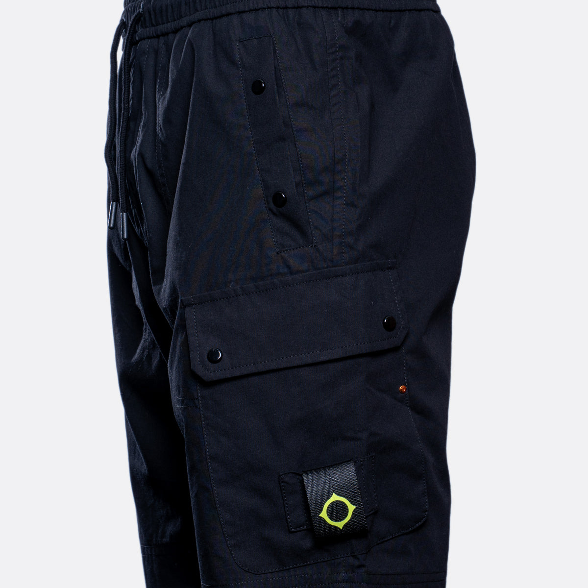 Close-up of black cargo shorts showing a side pocket with a black snap flap, a drawstring waist, and a small bright yellow-green circular logo patch on the lower leg. MA.Strum Shorts available at Sakks Menswear in Southampton.
