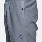 Close-up of gray athletic shorts with an elastic waistband, a vertical zip pocket with a black snap, and a blue embroidered geometric logo near the lower left leg. MA.Strum Swimwear available at Sakks Menswear in Southampton.