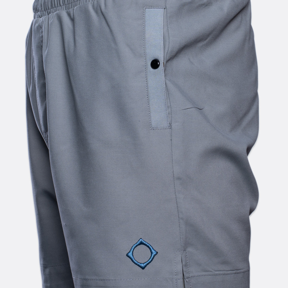 Close-up of gray athletic shorts with an elastic waistband, a vertical zip pocket with a black snap, and a blue embroidered geometric logo near the lower left leg. MA.Strum Swimwear available at Sakks Menswear in Southampton.