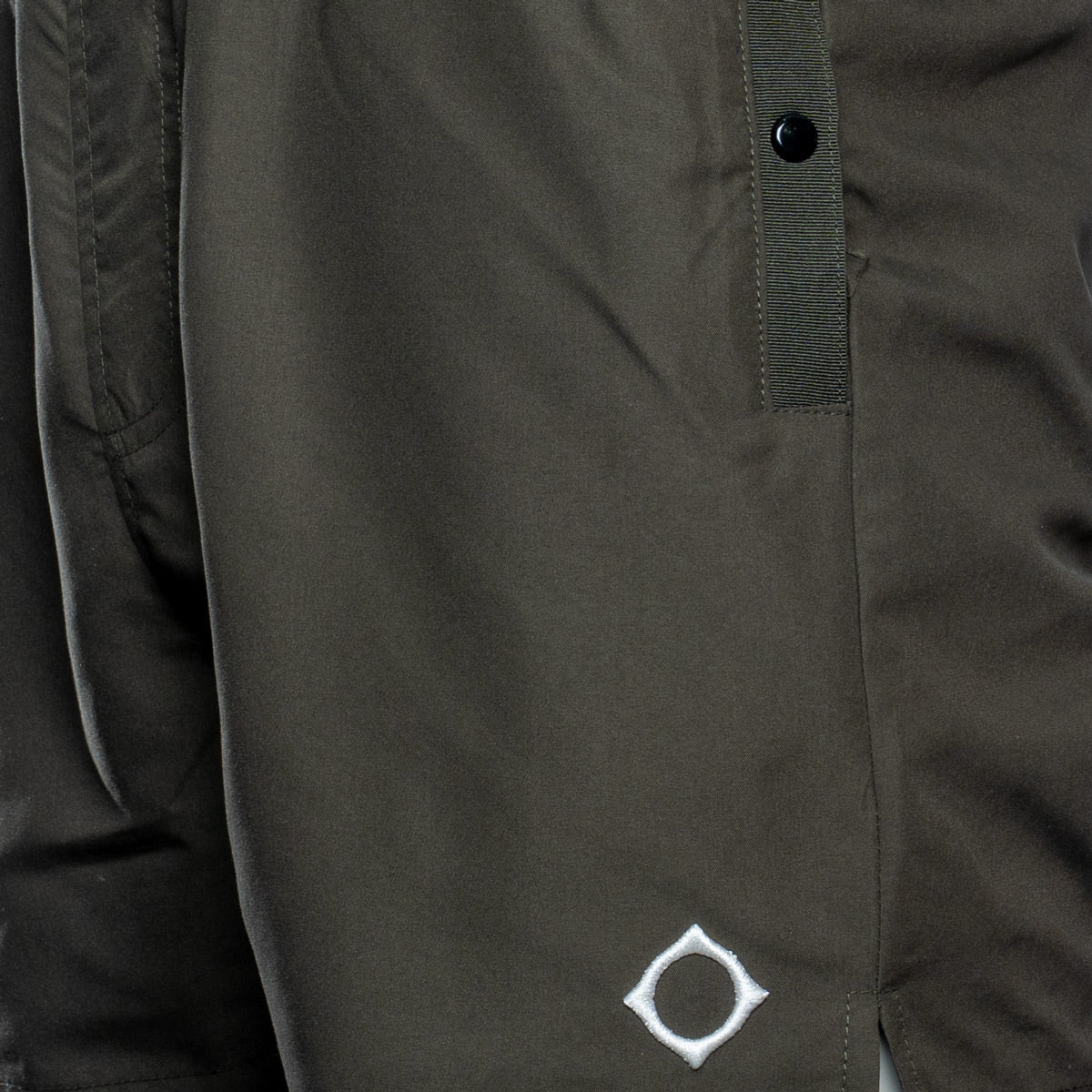 Close-up of dark olive green board shorts featuring a white embroidered diamond logo with a black center on the lower leg and a black snap button closure on a vertical strap. MA.Strum Swimwear available at Sakks Menswear in Southampton.