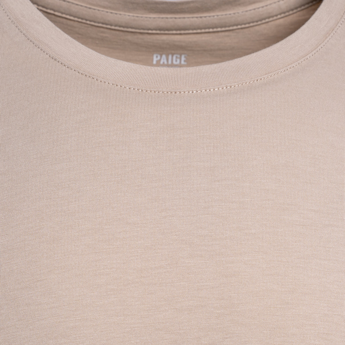 Close-up of a beige crewneck t-shirt with the brand name 'PAIGE' printed inside the collar. PAIGE T-Shirt available at Sakks Menswear in Southampton.