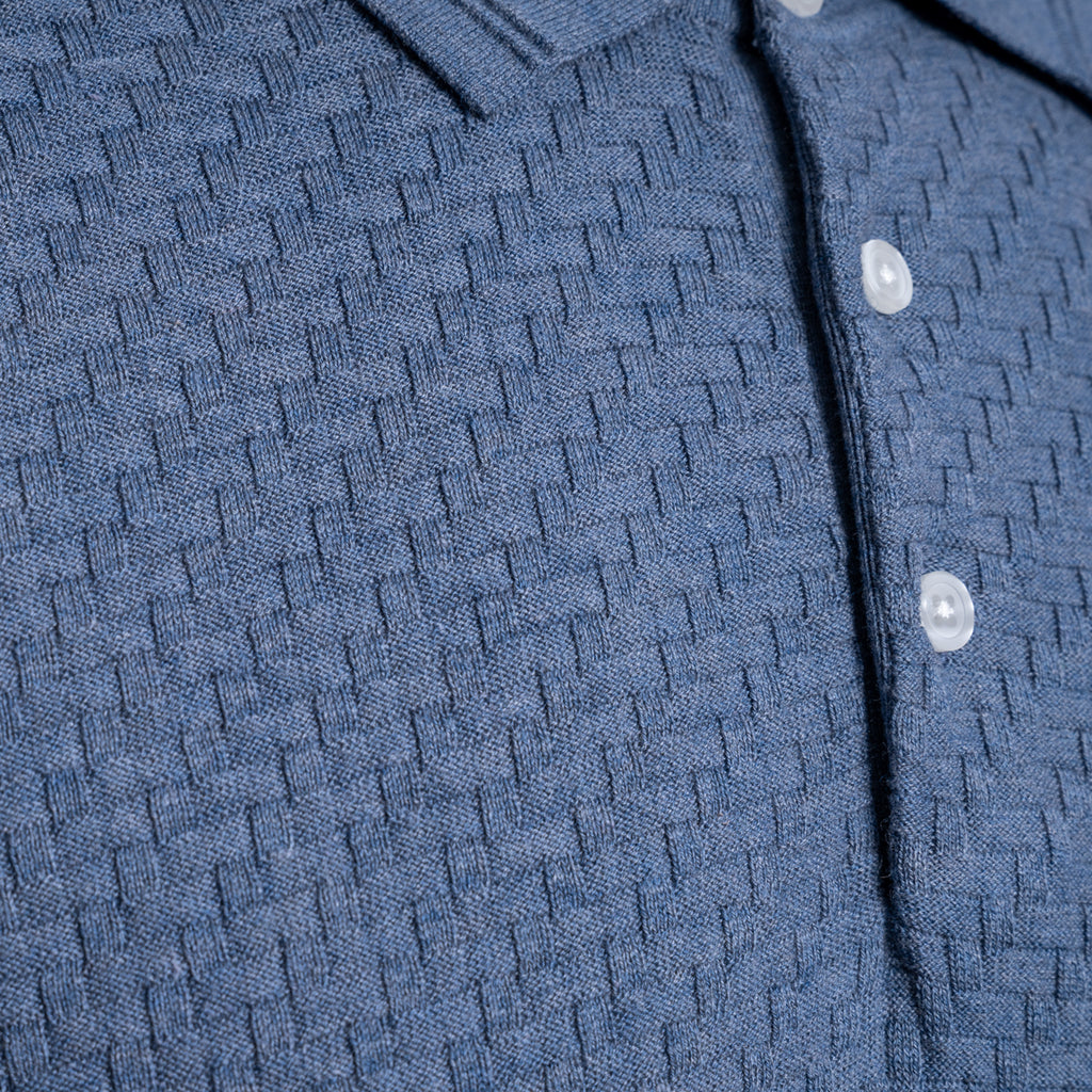 Close-up of a textured blue knit polo shirt with a buttoned collar and white buttons. PAIGE Polo available at Sakks Menswear in Southampton.
