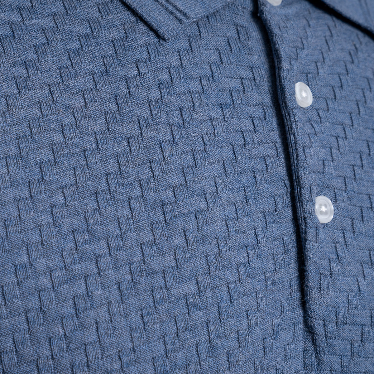 Close-up of a textured blue knit polo shirt with a buttoned collar and white buttons. PAIGE Polo available at Sakks Menswear in Southampton.