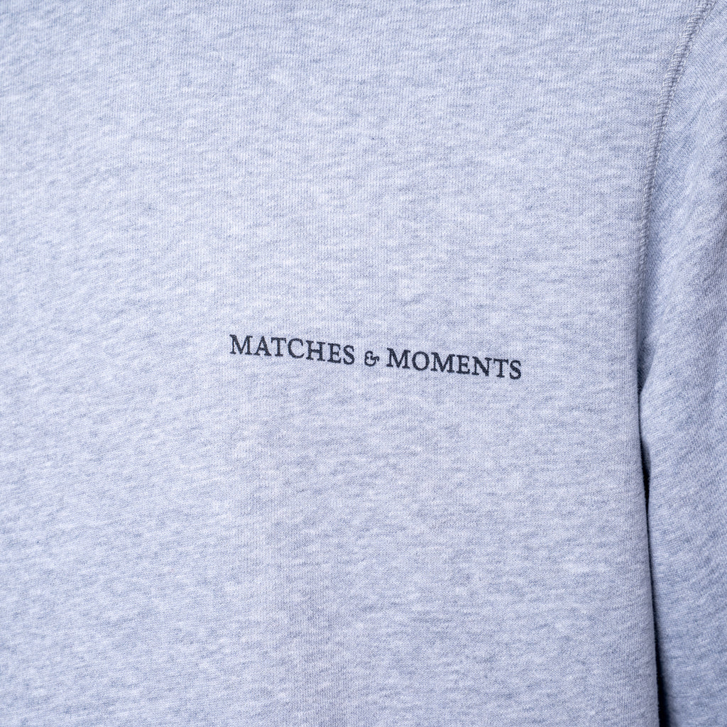 Close-up of a light heather gray sweatshirt with the black embroidered text 'MATCHES & MOMENTS' on the chest. PAIGE Sweatshirt available at Sakks Menswear in Southampton.
