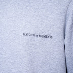 Close-up of a light heather gray sweatshirt with the black embroidered text 'MATCHES & MOMENTS' on the chest. PAIGE Sweatshirt available at Sakks Menswear in Southampton.