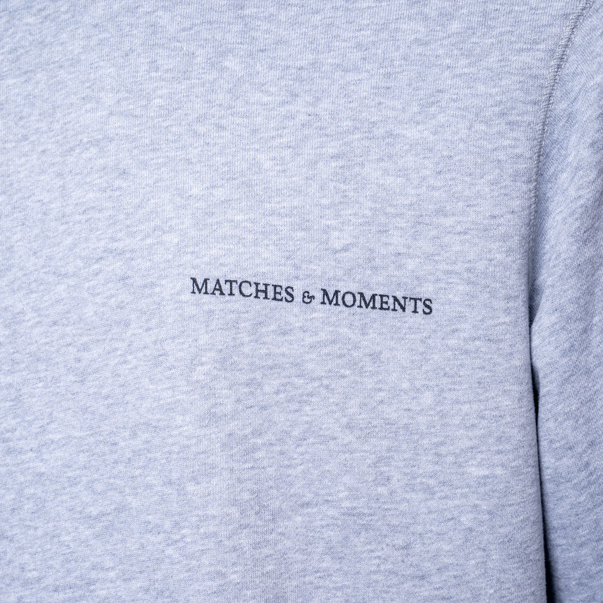 Close-up of a light heather gray sweatshirt with the black embroidered text 'MATCHES & MOMENTS' on the chest. PAIGE Sweatshirt available at Sakks Menswear in Southampton.