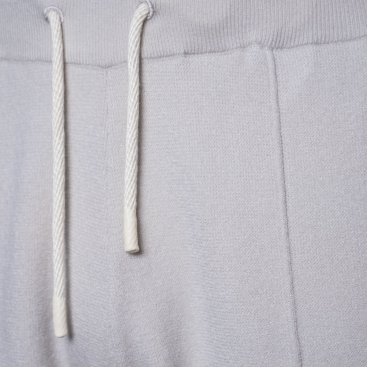 Close-up of a light gray fabric garment showing the ribbed waistband and two white drawstrings. PAIGE Sweat Shorts available at Sakks Menswear n Southampton.