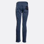A pair of slim-fit blue jeans shown from the back, featuring a black brand patch on the right back pocket, set against a plain light gray background. PAIGE Jeans available at Sakks Menswear in Southampton.