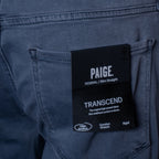 Close-up of the back pocket of a pair of Paige jeans with a black hangtag showing the brand name, 'PAIGE', and the model name 'TRANSCEND'. PAIGE Jeans available at Sakks Menswear in Southampton.
