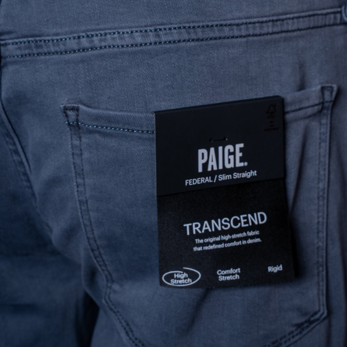 Close-up of the back pocket of a pair of Paige jeans with a black hangtag showing the brand name, 'PAIGE', and the model name 'TRANSCEND'. PAIGE Jeans available at Sakks Menswear in Southampton.