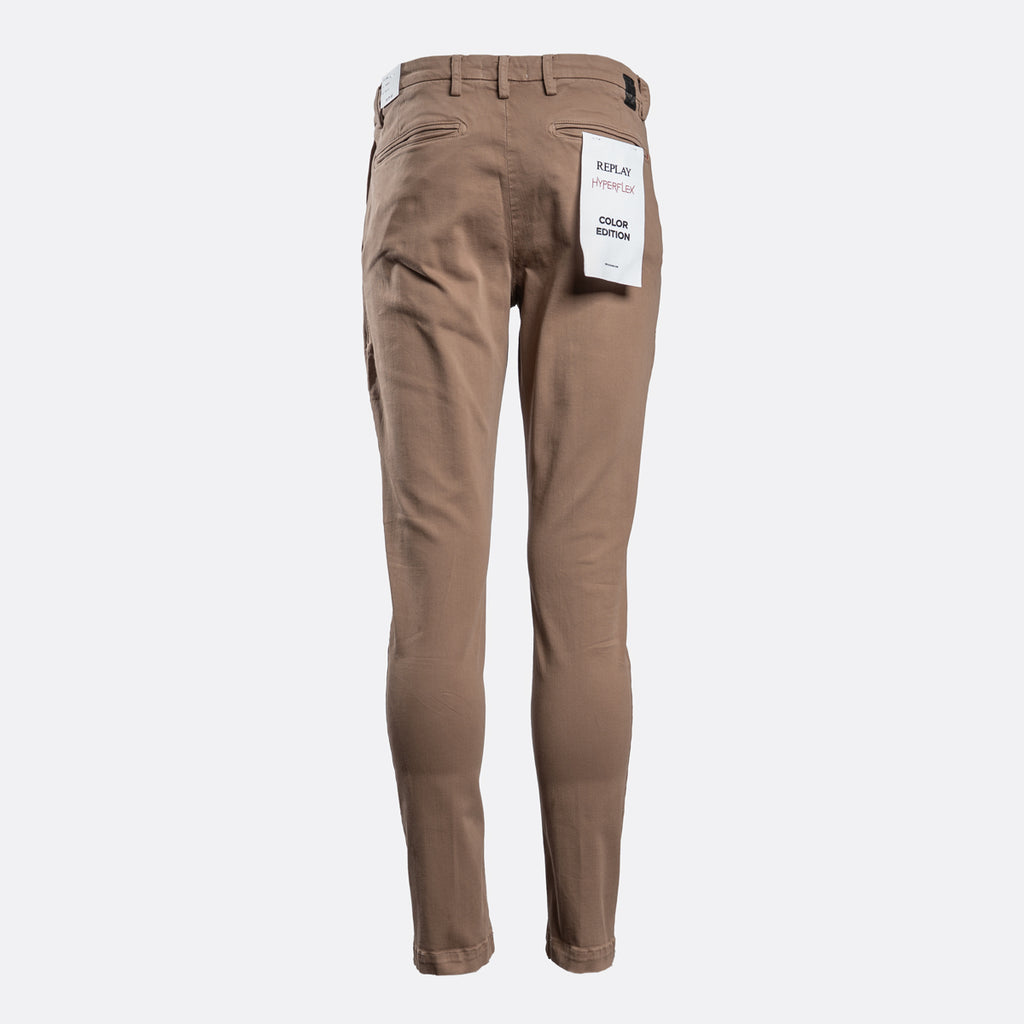 A pair of slim-fit brown pants shown from the back, featuring a white paper tag attached to the back pocket, set against a plain light gray background. Replay Chino available at Sakks Menswear in Southampton.