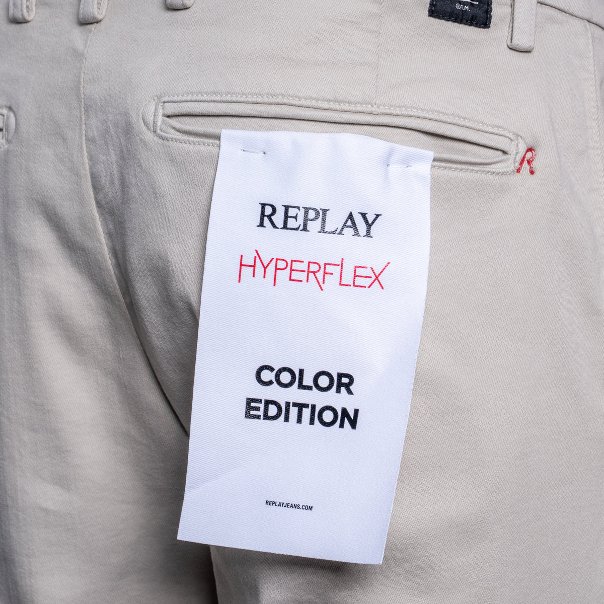 Close-up of beige Replay Hyperflex pants with a white paper hangtag reading 'REPLAY HYPERFLEX COLOR EDITION' attached near the back pocket. Replay Chinos available at Sakks Menswear in Southampton.