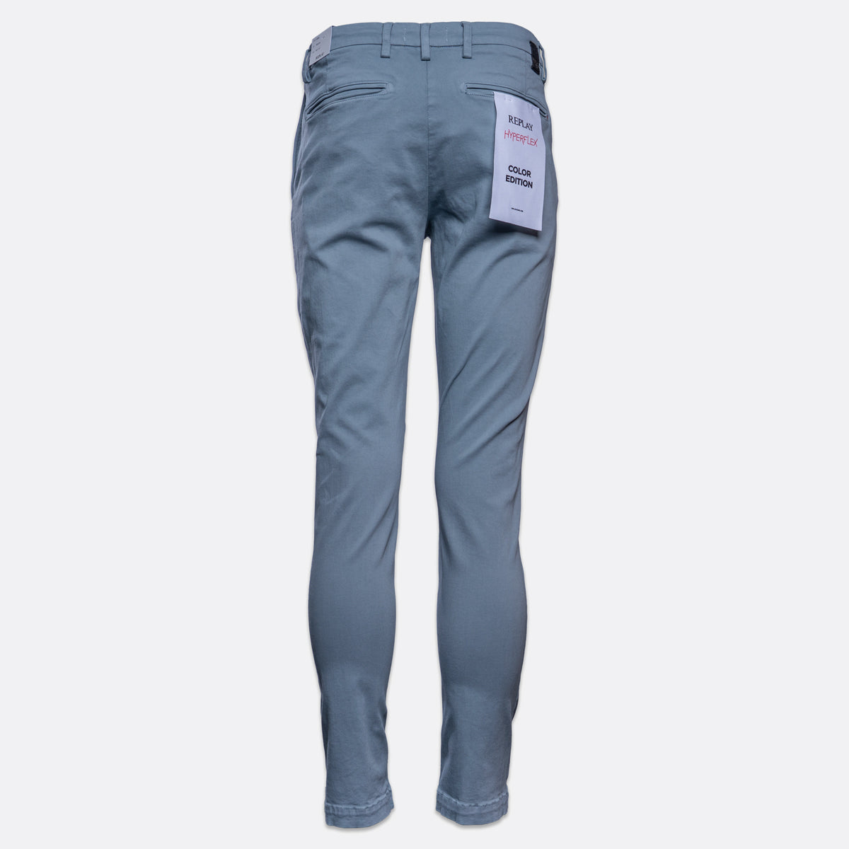 A pair of slim-fit blue jeans shown from the back, featuring a white paper tag attached to the back pocket, set against a plain light gray background. Replay Chinos available at Sakks Menswear in Southampton.