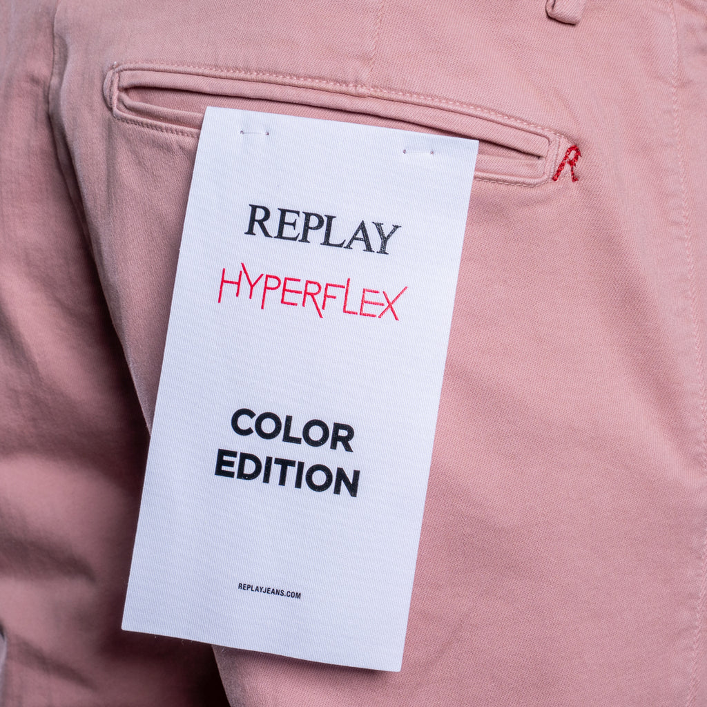 A close-up of dusty pink trousers showing the back pocket with a white paper tag that reads 'REPLAY HYPERFLEX COLOR EDITION'. Replay Chinos available at Sakks Menswear in Southampton.