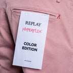 A close-up of dusty pink trousers showing the back pocket with a white paper tag that reads 'REPLAY HYPERFLEX COLOR EDITION'. Replay Chinos available at Sakks Menswear in Southampton.
