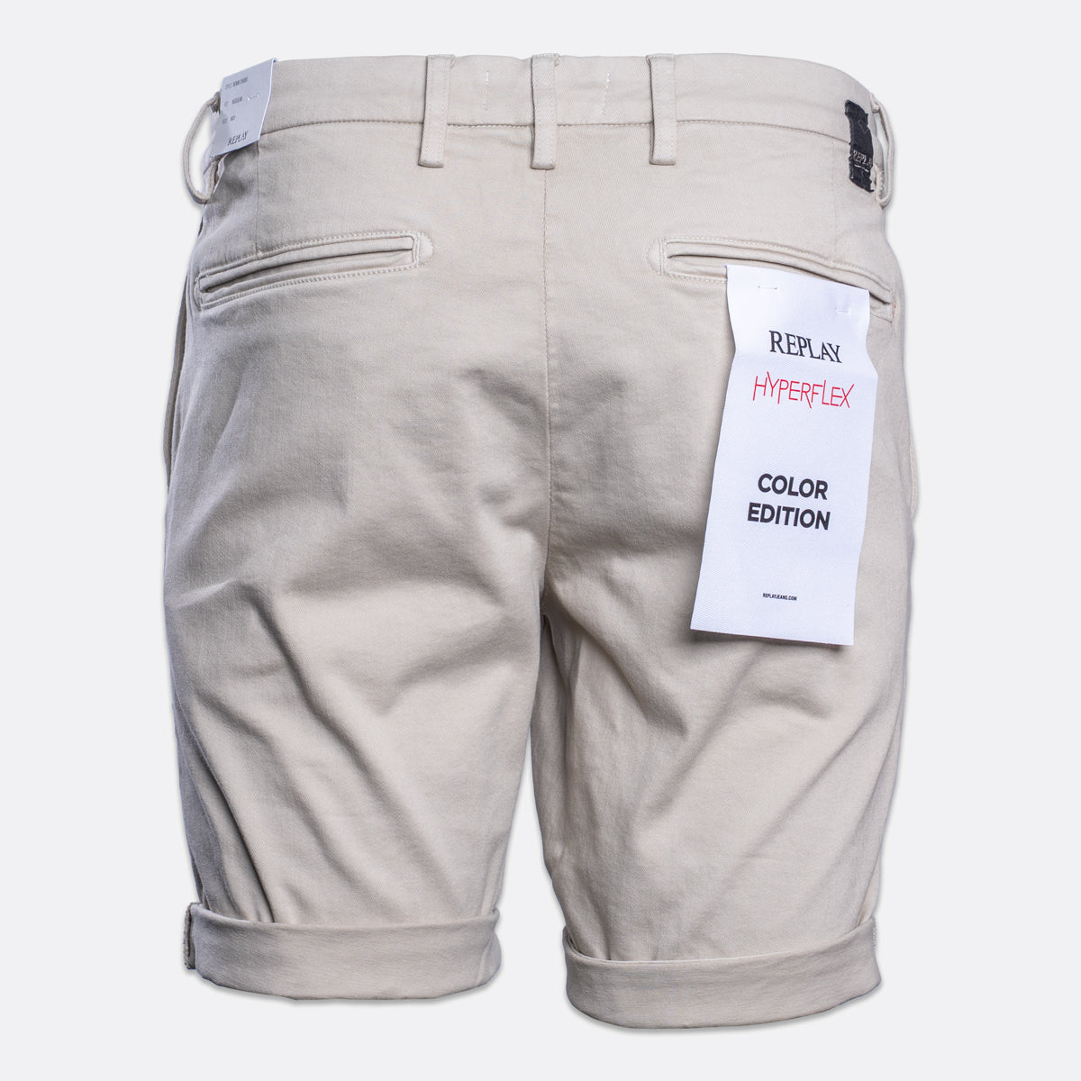 Back view of light beige chino shorts with rolled cuffs, featuring a white paper tag on the right pocket that reads 'REPLAY HYPERFLEX COLOR EDITION'. Replay Shorts available at Sakks Menswear in Southampton.