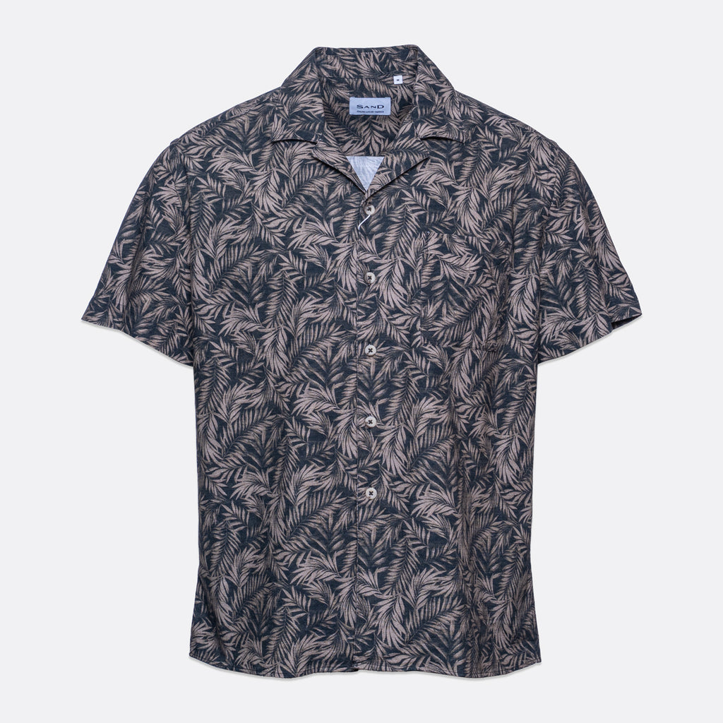 A short-sleeve button-up camp shirt by Branded Play, featuring an all-over palm leaf pattern in dark navy and light beige, displayed on a clean light gray background. Sand Copenhagen Short Sleeve Shirt available at Sakks Menswear in Southampton.
