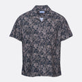 A short-sleeve button-up camp shirt by Branded Play, featuring an all-over palm leaf pattern in dark navy and light beige, displayed on a clean light gray background. Sand Copenhagen Short Sleeve Shirt available at Sakks Menswear in Southampton.