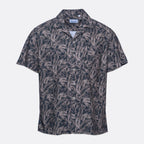 A short-sleeve button-up camp shirt by Branded Play, featuring an all-over palm leaf pattern in dark navy and light beige, displayed on a clean light gray background. Sand Copenhagen Short Sleeve Shirt available at Sakks Menswear in Southampton.