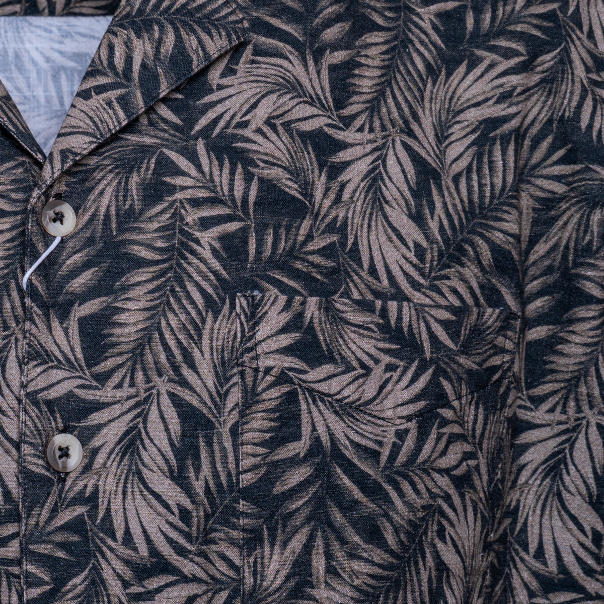 Close-up of a button-front shirt with an allover tropical leaf print in dark navy and muted taupe-gray. Sand Copenhagen Short Sleeve Shirt available at Sakks Menswear in Southampton.