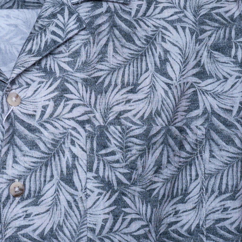 Close-up of a button-front shirt with a gray and white tropical palm leaf pattern, showing part of the collar and two light-colored buttons. Sand Copenhagen Short Sleeve Shirt available at Sakks Menswear in Southampton.