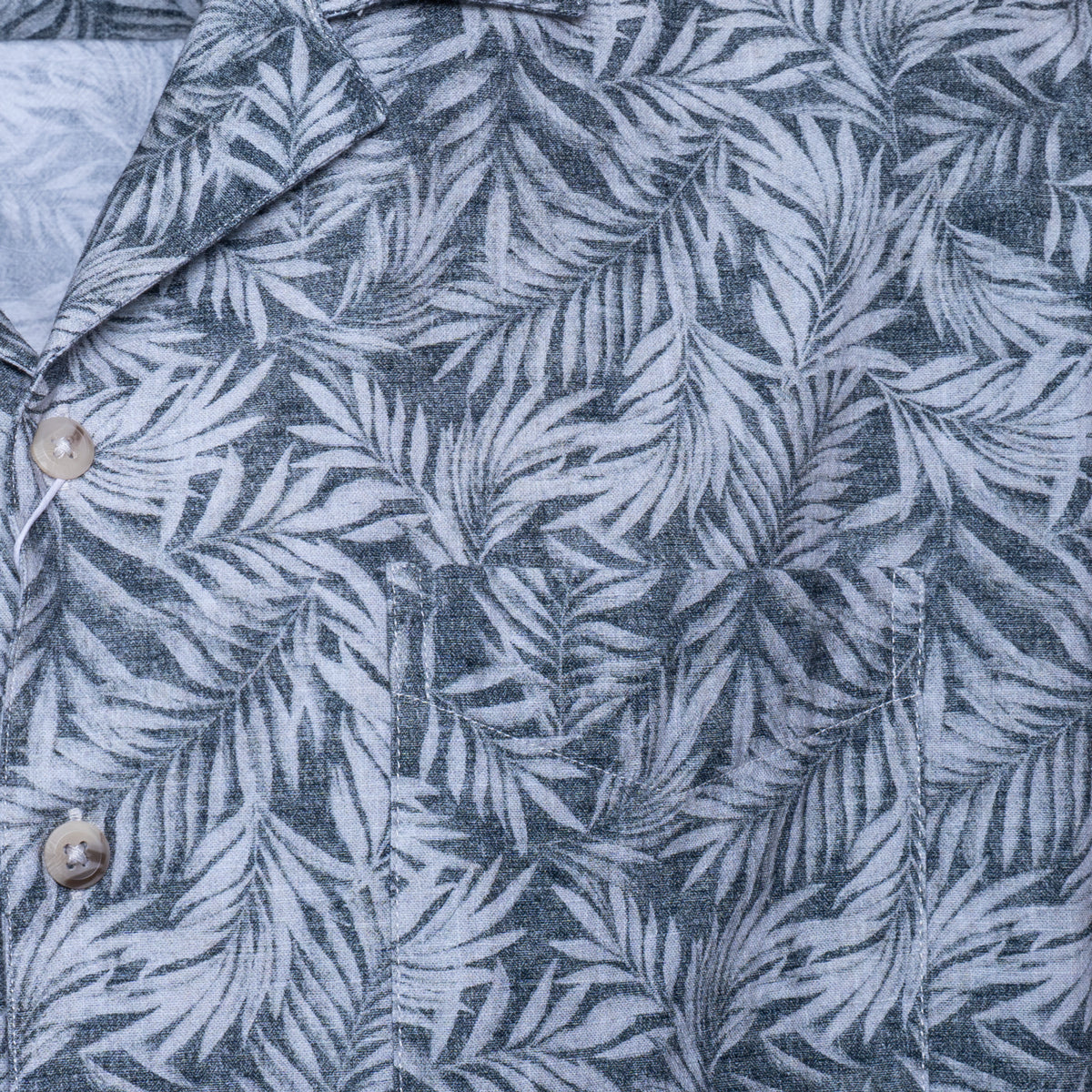 Close-up of a button-front shirt with a gray and white tropical palm leaf pattern, showing part of the collar and two light-colored buttons. Sand Copenhagen Short Sleeve Shirt available at Sakks Menswear in Southampton.