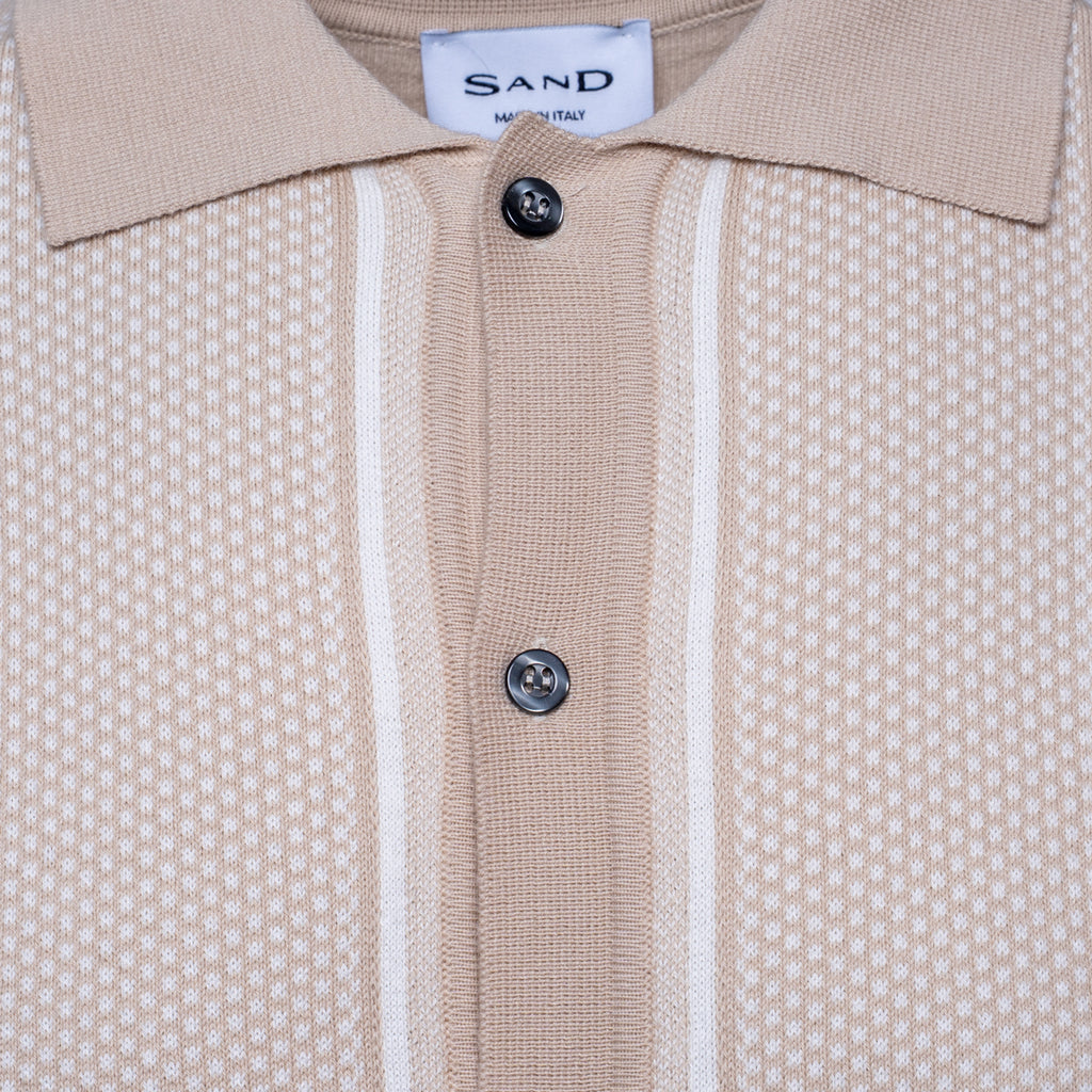 Close-up of a beige knit polo shirt with a textured micro-dot pattern, featuring two vertical white stripes down the center, a white collar, and two dark buttons. The brand label 'SAND' is visible inside the collar. Sand Copenhagen Polo available at Sakks Menswear in Southampton.