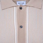 Close-up of a beige knit polo shirt with a textured micro-dot pattern, featuring two vertical white stripes down the center, a white collar, and two dark buttons. The brand label 'SAND' is visible inside the collar. Sand Copenhagen Polo available at Sakks Menswear in Southampton.