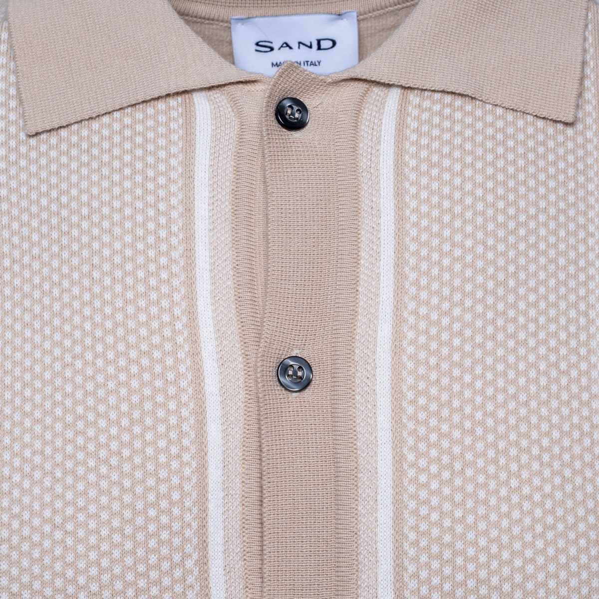 Close-up of a beige knit polo shirt with a textured micro-dot pattern, featuring two vertical white stripes down the center, a white collar, and two dark buttons. The brand label 'SAND' is visible inside the collar. Sand Copenhagen Polo available at Sakks Menswear in Southampton.
