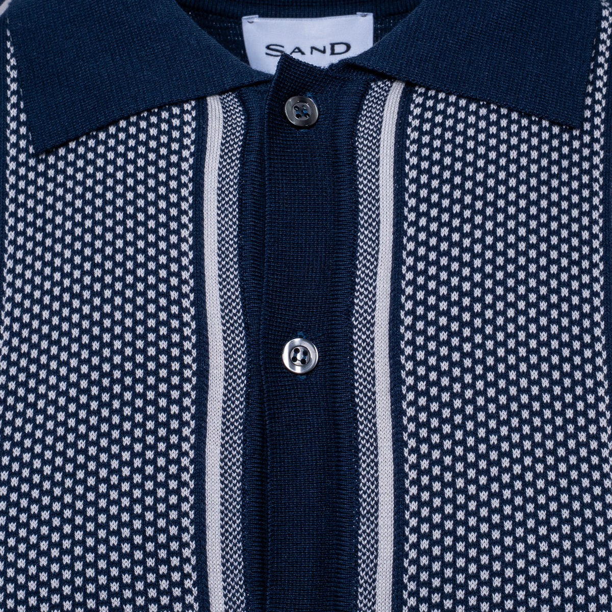 Close-up of a knit polo shirt collar and button placket. The shirt features a dark navy blue collar and center placket, with textured side panels in a navy and white micro-pattern, separated by thin white vertical stripes. Sand Copenhagen Polo available at Sakks Menswear in Southampton.