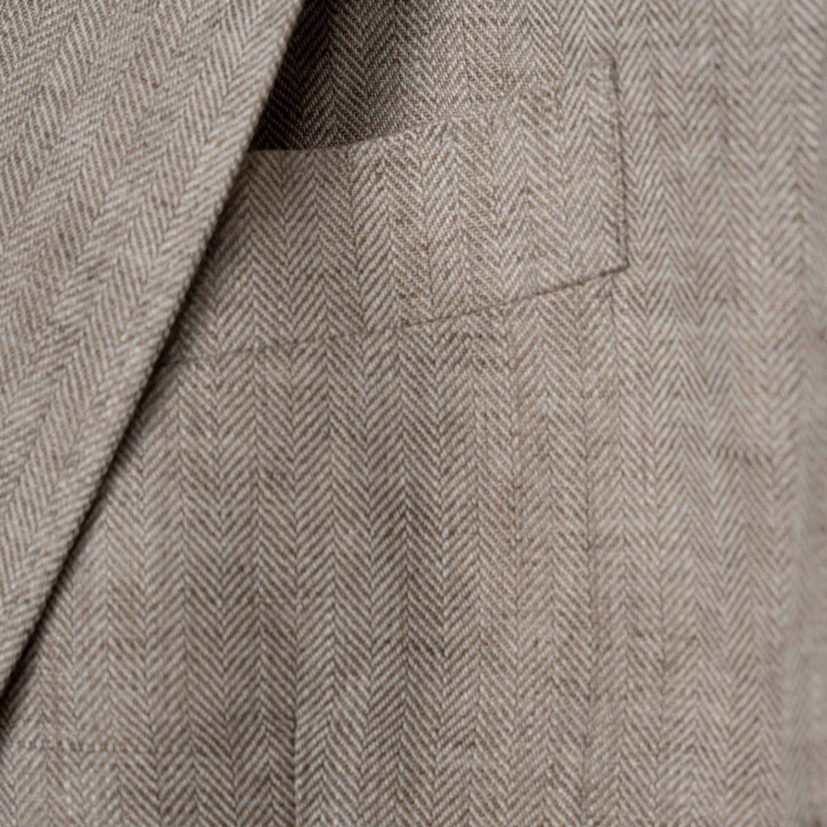 Close-up of a beige and brown herringbone tweed fabric with a pocket and lapel detail. Sand Copenhagen Blazer available at Sakks Menswear in Southampton.