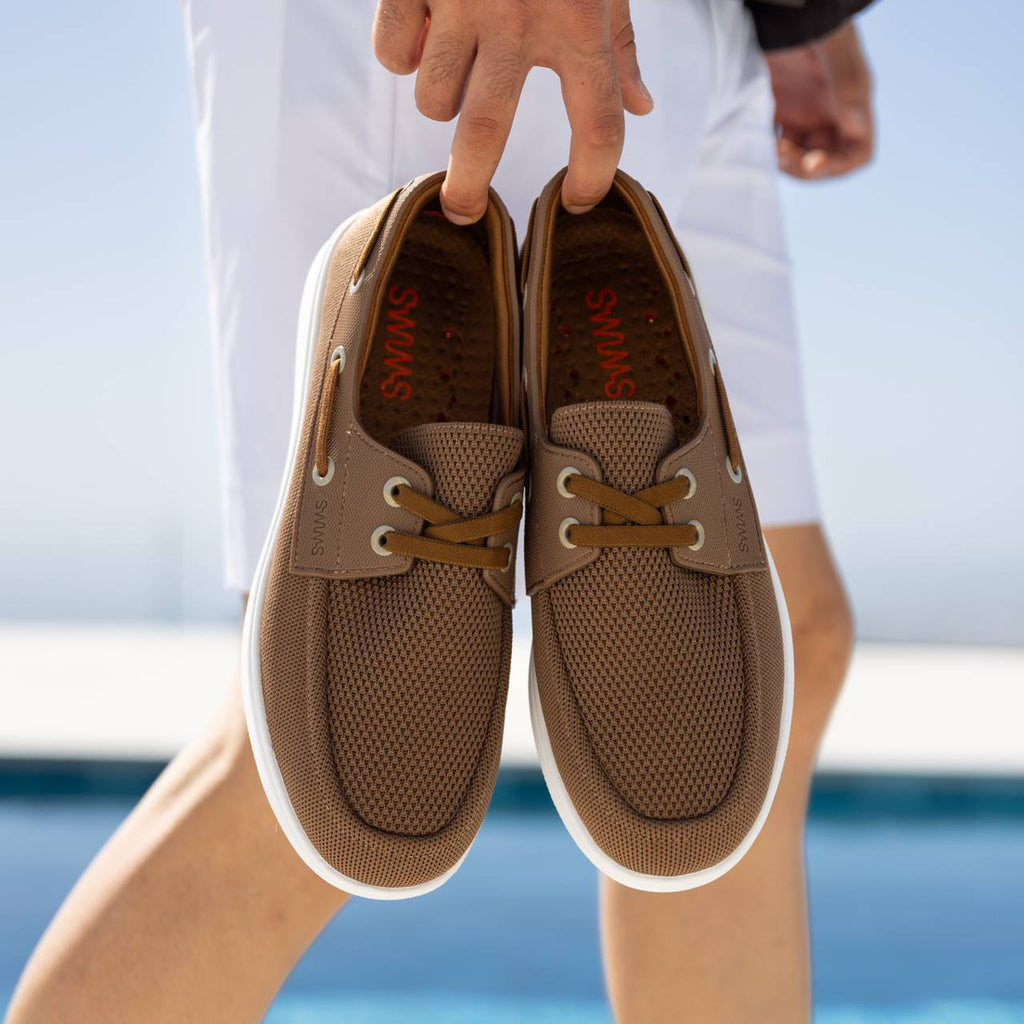 A person holding a pair of brown mesh boat shoes with white soles, photographed outdoors near a pool with the person's legs and hands visible. SWIMS Boat Shoes available at Sakks Menswear in Southampton.
