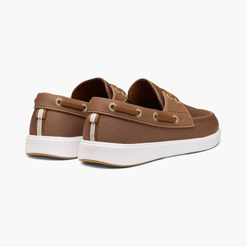 A pair of brown boat shoes with white soles, shown from a three-quarters rear angle on a light gray background. SWIMS Boat Shoes available at Sakks Menswear in Southampton.