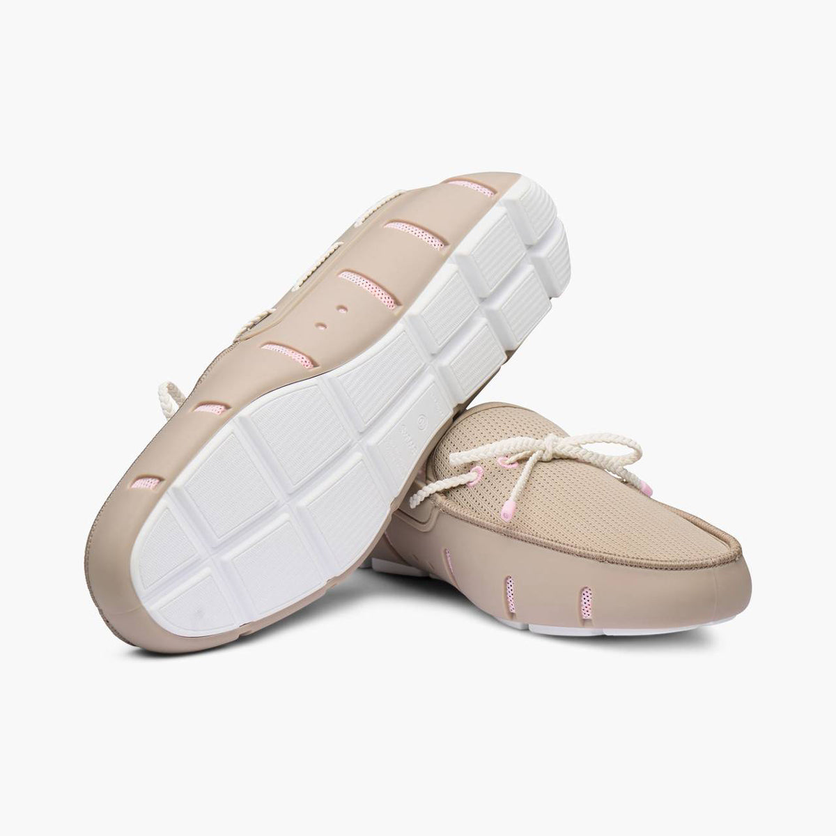 A pair of slip-on boat shoes in beige with white laces and light pink accents, shown from the side and bottom against a light gray background. SWIMS Loafers available at Sakks Menswear in Southampton.