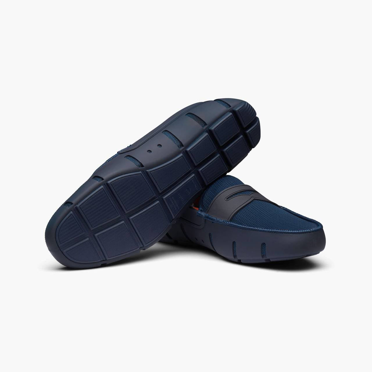 A pair of navy blue slip-on loafers shown on a clean white background, with one shoe angled to display the textured outsole and the other showing the side profile. SWIMS Loafers available at Sakks Menswear in Southampton.