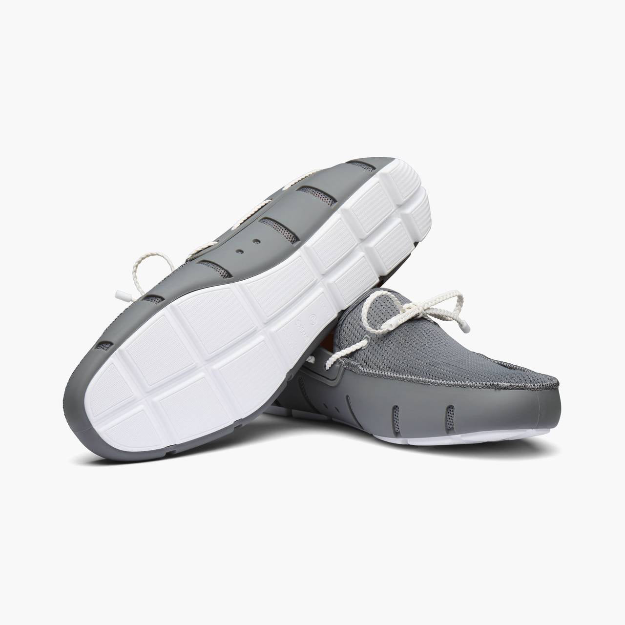 A pair of gray slip-on boat shoes with white laces, shown from a three-quarters perspective to display both the side profile and the textured white outsole. SWIMS Loafers available at Sakks Menswear in Southampton.