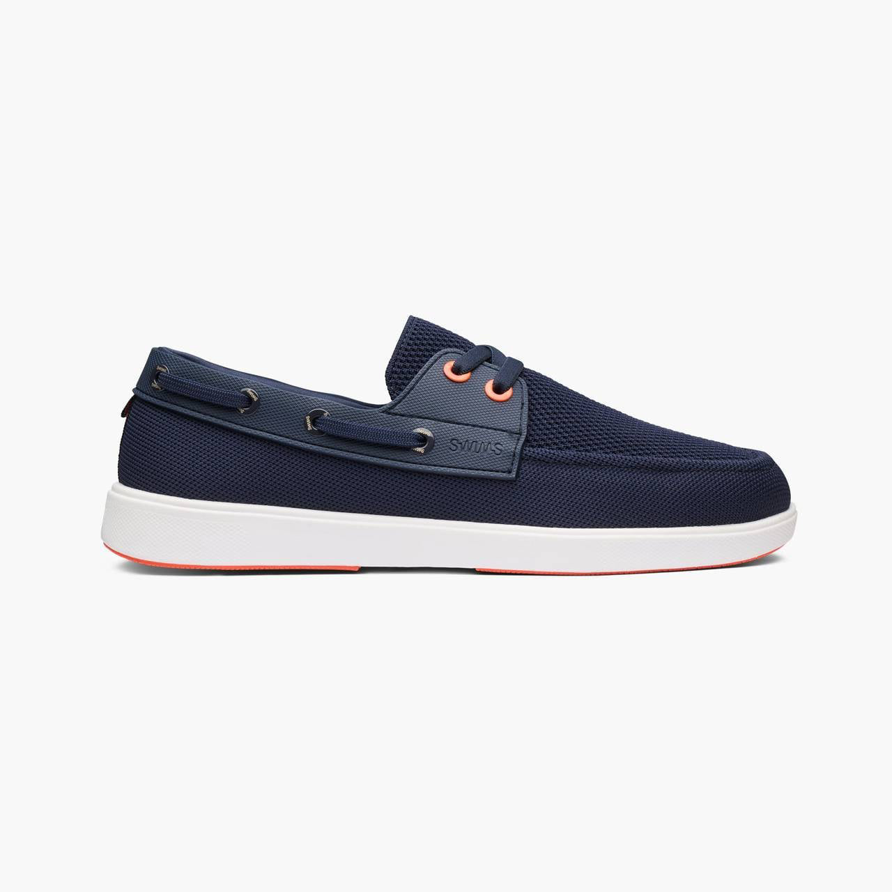 A navy blue boat shoe with a white sole and orange eyelets, shown in a side profile on a light gray background. SWIMS Boat Shoes available at Sakks Menswear in Southampton.