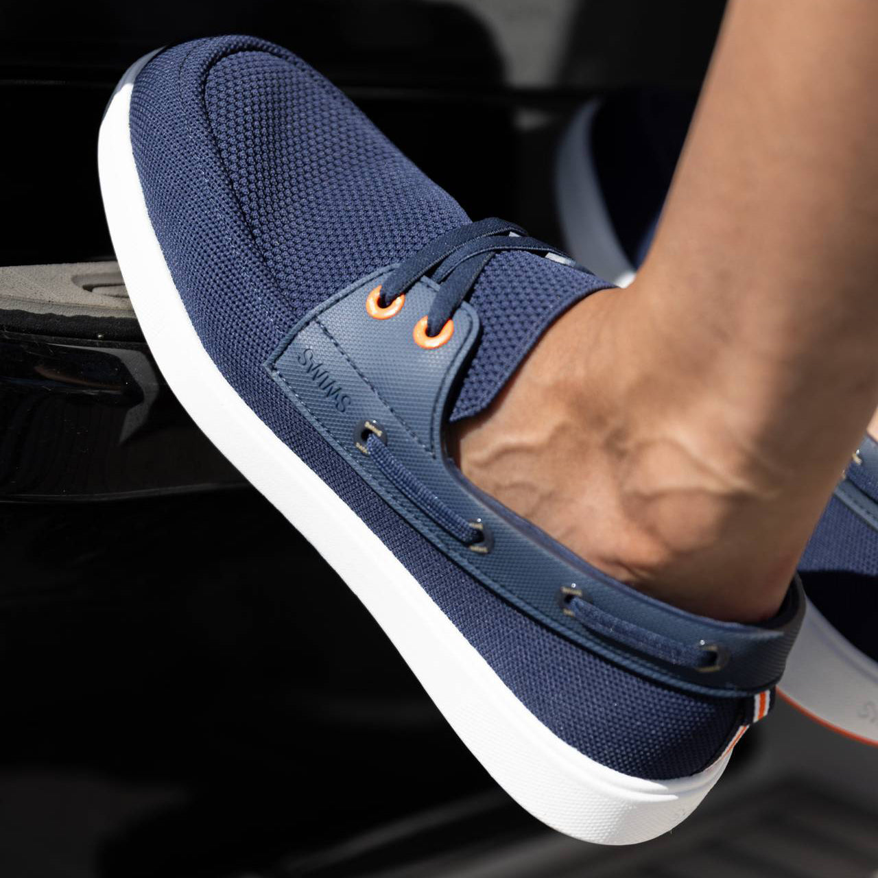 A close-up of a person's foot wearing a navy blue mesh slip-on boat shoe with a white sole, resting on a dark surface. SWIMS Boat Shoes available at Sakks Menswear in Southampton.