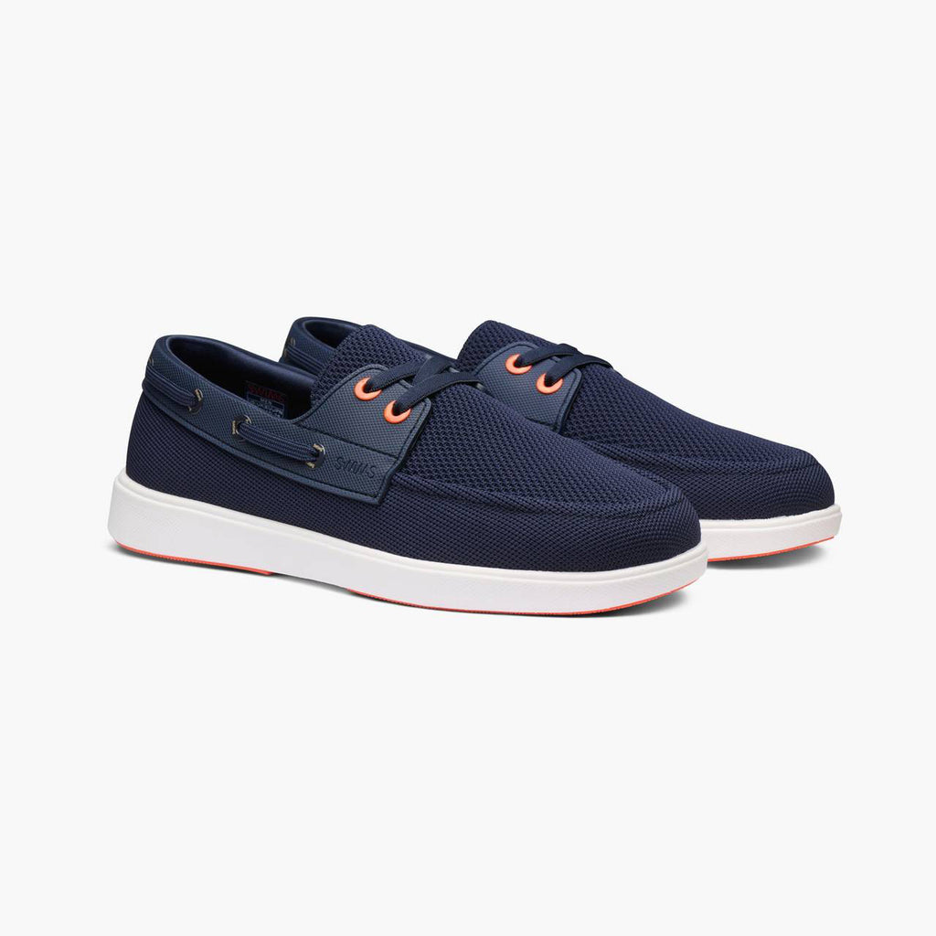 A pair of navy blue mesh boat shoes with white soles and orange eyelets, shown on a light gray background. SWIMS Boat Shoes available at Sakks Menswear in Southampton.
