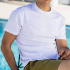 A man wearing a plain white short-sleeve crewneck t-shirt and olive green shorts, sitting by a swimming pool. SWIMS T-Shirt available at Sakks Menswear in Southampton.
