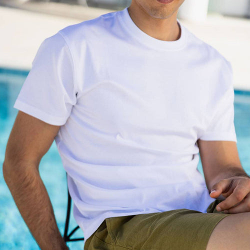A man wearing a plain white short-sleeve crewneck t-shirt and olive green shorts, sitting by a swimming pool. SWIMS T-Shirt available at Sakks Menswear in Southampton.