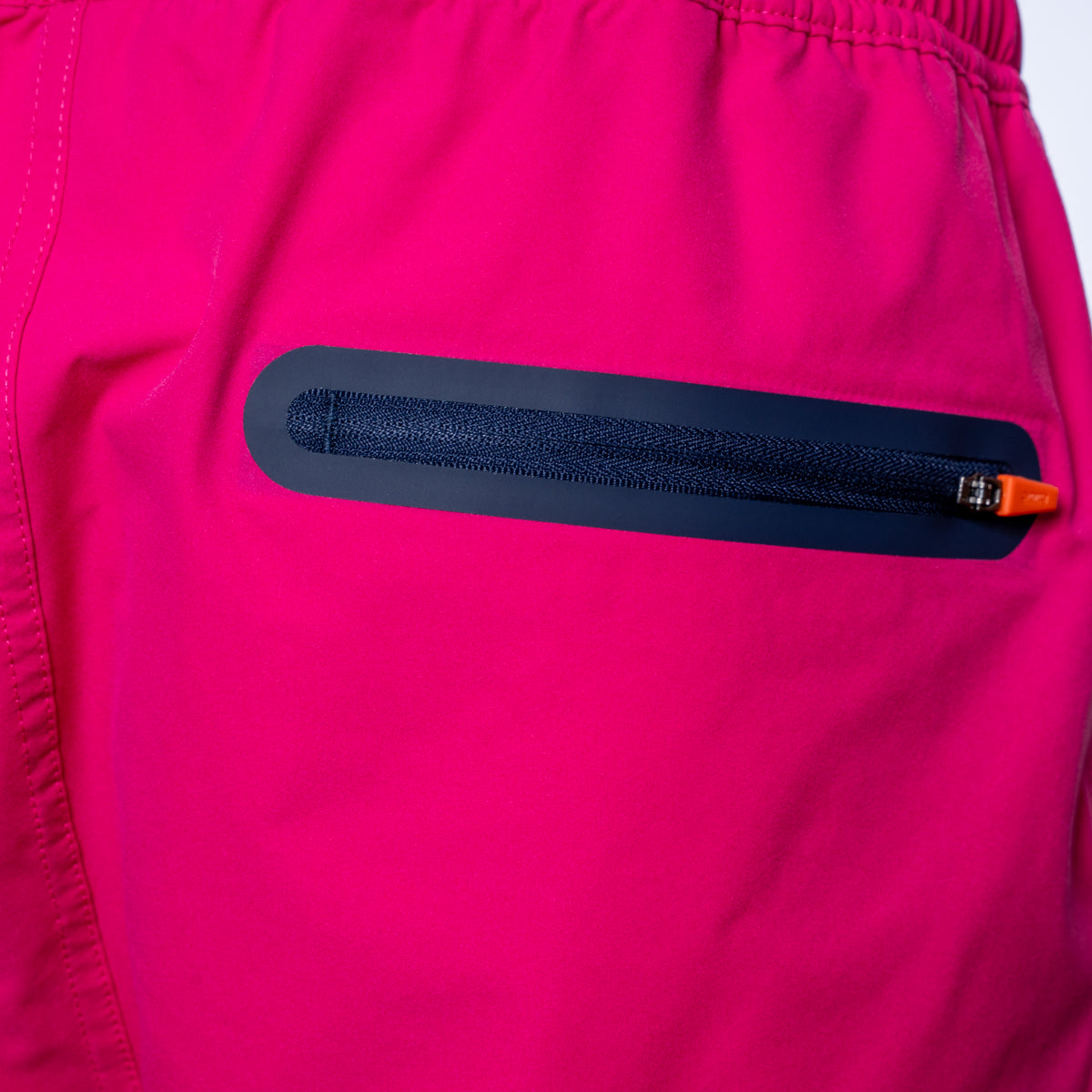 Close-up of a bright pink garment featuring a horizontal black zippered pocket with a small orange zipper pull. SWIMS Swimshorts available at Sakks Menswear in Southampton.