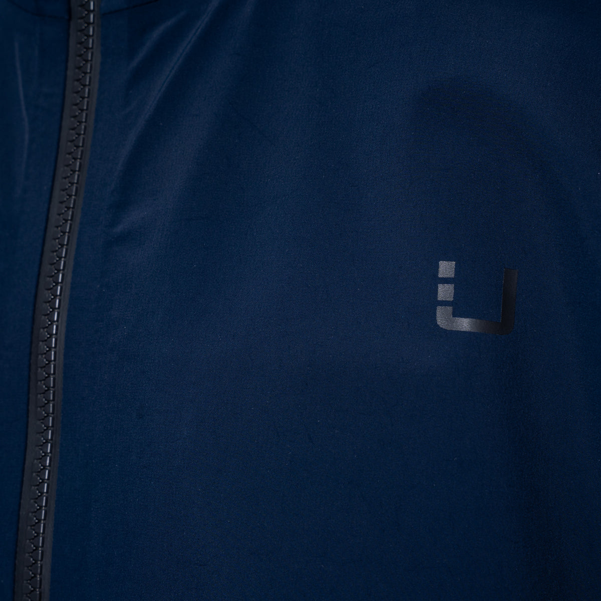 Close-up of a dark navy blue jacket with a black zipper on the left and a small dark gray stylized 'U' logo on the chest. UBR Jacket available at Sakks Menswear in Southampton.