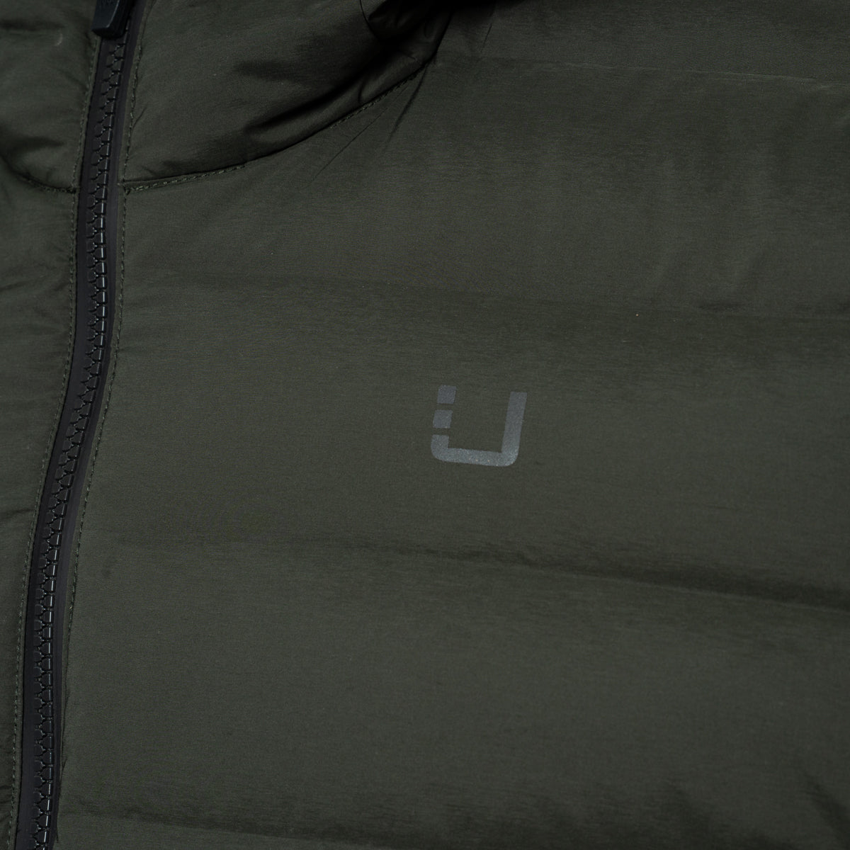 Close-up of an olive green puffer jacket with a dark zipper and a small gray stylized logo on the chest. UBR Jacket available at Sakks Menswear in Southampton.