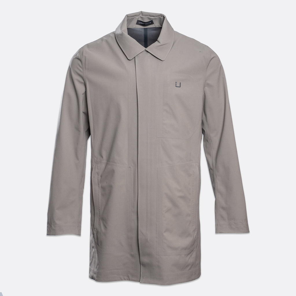 A light gray utility jacket with a pointed collar, long sleeves, and a small square logo patch on the left chest, displayed against a light gray background. UBR Jacket available at Sakks Menswear in Southampton.